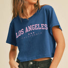 A woman wearing a cropped blue t-shirt with 'LOS ANGELES CALIFORNIA' printed in pink and white across the chest, paired with dark gray jeans, set against a plain beige background.