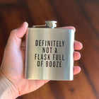 Hand holding a stainless steel flask with text 'DEFINITELY NOT A FLASK FULL OF BOOZE' against a blurred background.