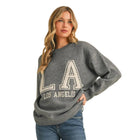 A woman modeling a gray crewneck sweater with 'LA LOS ANGELES' printed in white varsity lettering, paired with blue skinny jeans and white sneakers, against a solid beige studio background.