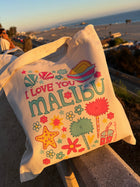 A cream canvas tote bag with a colorful 'I LOVE YOU MALIBU' beach graphic, resting on a stone railing overlooking a scenic coastal view at sunset.