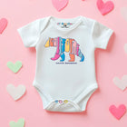 A white short-sleeve baby bodysuit laid flat on a pink background with pastel heart-shaped decorations. The bodysuit has a colorful bear graphic with the word 'California' and 'Santa Barbara' printed on the front.