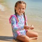 Young girl in a colorful swimsuit sitting on a beach with clear water and sky.