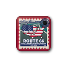 A square pinback button badge featuring a vintage-style Route 66 travel stamp design. The badge has a red border, a map of the United States filled with the American flag, and text reading 'Greetings From', 'ROUTE 66', 'THE AMERICAN HIGHWAY', and 'EST.