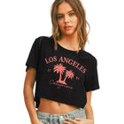 A woman modeling a black cropped graphic t-shirt with pink palm tree and 'Los Angeles California' text, paired with light-wash denim shorts, against a solid beige background.