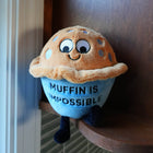 Plush toy resembling a muffin with text 'Muffin is Impossible' held against a wooden surface.