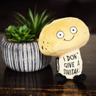 Plush toy resembling a mushroom with a humorous message next to a potted plant on a wooden surface.
