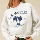 A woman wearing a cream-colored crewneck sweatshirt with a navy blue 'Los Angeles California' palm tree graphic, paired with a beige cap and black pants, set against a solid beige background.