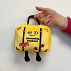 Plush toy suitcase labeled 'Emotional Baggage' held by a hand on a white background