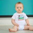 Baby wearing a onesie with a California design sitting on a light-colored floor against a teal wall.