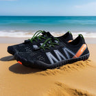 A pair of black water shoes with green laces and orange accents, resting on a sandy beach with the ocean and blue sky in the background.
