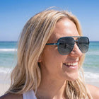 A smiling blonde woman wearing dark aviator sunglasses stands on a sandy beach with the ocean and blue sky in the background.