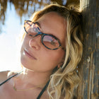 Woman wearing glasses leaning against a wooden wall with sunlight filtering through.