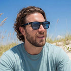 Man wearing sunglasses sitting on a sandy beach with tall grasses.