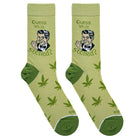 A pair of light olive green crew socks with dark green heels and toes, featuring a cartoon illustration of a man and the text 'Guess Who's Stoned?' along with scattered green cannabis leaf prints.