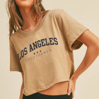 A woman wearing a cropped beige t-shirt with 'LOS ANGELES CALIFORNIA' printed in navy blue across the chest, paired with black bottoms, set against a plain beige background.