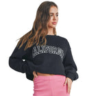 A woman modeling a black cropped sweatshirt with 'CALIFORNIA' in white arched letters, paired with bright pink biker shorts, white socks, and white sneakers, standing against a light gray wall.