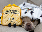 Dog lying on a couch with a plush toy resembling a slice of cheese labeled 'It ain't easy being cheesy'.