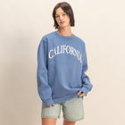 Person wearing a blue 'California' sweatshirt with denim shorts on a beige background.