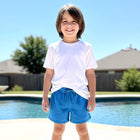 A young boy stands barefoot on a concrete pool deck in front of a swimming pool and house, wearing a white short-sleeve t-shirt and bright blue drawstring shorts.