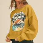 A woman wearing a mustard yellow crewneck sweatshirt with a colorful retro 'Santa Monica California' beach graphic, paired with black jeans and a white cap, against a solid beige background.