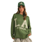 A woman modeling an oversized olive green sweatshirt with distressed white 'LA Los Angeles' lettering, paired with striped shorts, a green 'LA' baseball cap, white socks, and gray sneakers, against a solid beige studio background.