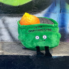 Green plush toy with a face and text, sitting on a concrete surface with graffiti in the background.