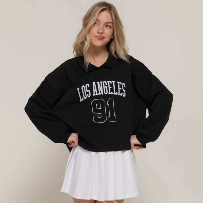 Person wearing a black 'Los Angeles 91' sweatshirt and white pleated skirt on a plain background