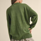 A woman shown from the back wearing an oversized olive green knit sweater over a pleated striped skirt, against a plain beige studio background.