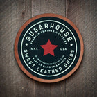 Round leather coaster with 'Sugarhouse Leather Goods' branding on a wooden surface