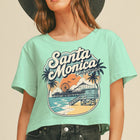 A woman wearing a mint green cropped t-shirt with a retro Santa Monica California graphic, paired with a wide-brimmed black hat and dark jeans, set against a solid beige background.