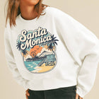 A woman wearing a white crewneck sweatshirt with a colorful 'Santa Monica California' beach graphic, paired with black jeans and a beige baseball cap, against a solid beige background.