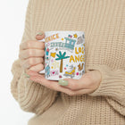 Model holding a mug with 'Venice Los Angeles' design, wearing a beige sweater.