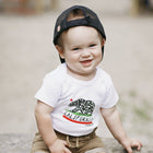 Baby wearing a white t-shirt with a California bear design and a black cap.