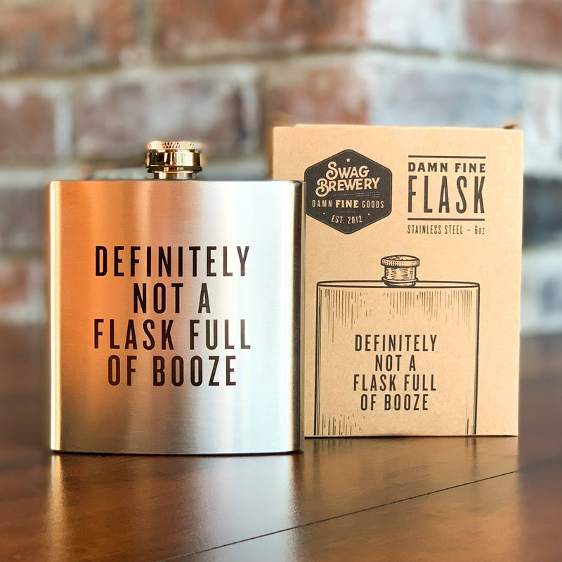 Metallic flask with humorous text next to its packaging on a wooden surface.