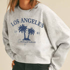 A woman wearing a light heather gray sweatshirt with a navy blue 'Los Angeles California' palm tree graphic, paired with a beige baseball cap and dark jeans, against a solid beige background.