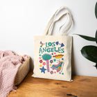 A cream canvas tote bag with a colorful 'Los Angeles' graphic print, styled on a wooden table next to a pink blanket and a green plant against a white wall.