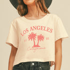 A woman wearing a cream-colored cropped t-shirt with a pink 'Los Angeles California' palm tree graphic, paired with dark jeans and a wide-brimmed black hat, set against a solid beige background.