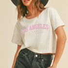 A woman wearing a cream-colored cropped t-shirt with pink 'Los Angeles' text, paired with a wide-brimmed dark gray hat and dark denim shorts, against a solid beige background.