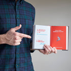 Person holding a book open with text and pointing at it, wearing a plaid shirt.