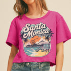 A woman wearing a bright pink cropped graphic T-shirt with a colorful Santa Monica, California beach scene print, paired with blue jeans against a plain beige background.