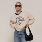 Person wearing a beige sweatshirt with text, blue jeans, and sunglasses on a plain background