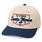 A structured baseball cap with a cream-colored crown and a navy blue brim. The front features an embroidered shield-shaped patch with the text 'CALIFORNIA Santa Monica 1908' and a palm tree in navy blue and red.