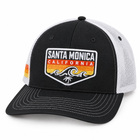 A black and white trucker hat featuring a prominent embroidered 'Santa Monica California' patch on the front. The cap has a black front panel and brim, white mesh back panels, and white contrast stitching.
