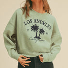 A woman wearing a light sage green crewneck sweatshirt with a black 'Los Angeles California' palm tree graphic, paired with black denim shorts and a beige hat, against a solid beige background.
