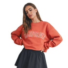 A woman modeling a coral orange sweatshirt with 'CALIFORNIA' printed across the chest, paired with a black pleated mini skirt, white socks, and white sneakers, standing against a light gray wall.