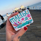 Hand holding a colorful 'Malibu' sticker with a beach scene on a wooden pier.