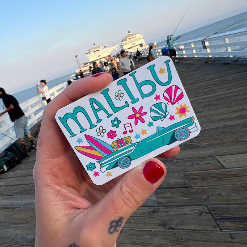 Hand holding a colorful 'Malibu' sticker with a beach scene on a wooden pier.