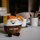 Plush toy resembling a coffee pod with 'POT HEAD' text on a desk with cups and pens.