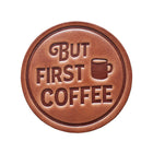 Round brown leather coaster with 'But First Coffee' text and coffee cup icon on a wooden surface.