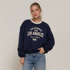 Woman wearing a navy sweatshirt with 'Los Angeles' text and blue jeans on a plain background
